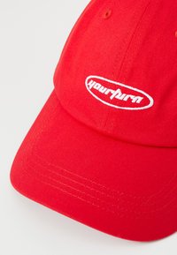 Rood katoenen baseballcap met een gebogen rand, met witte geborduurde tekst in een ovale vorm aan de voorkant. De tekst luidt "yourturn."