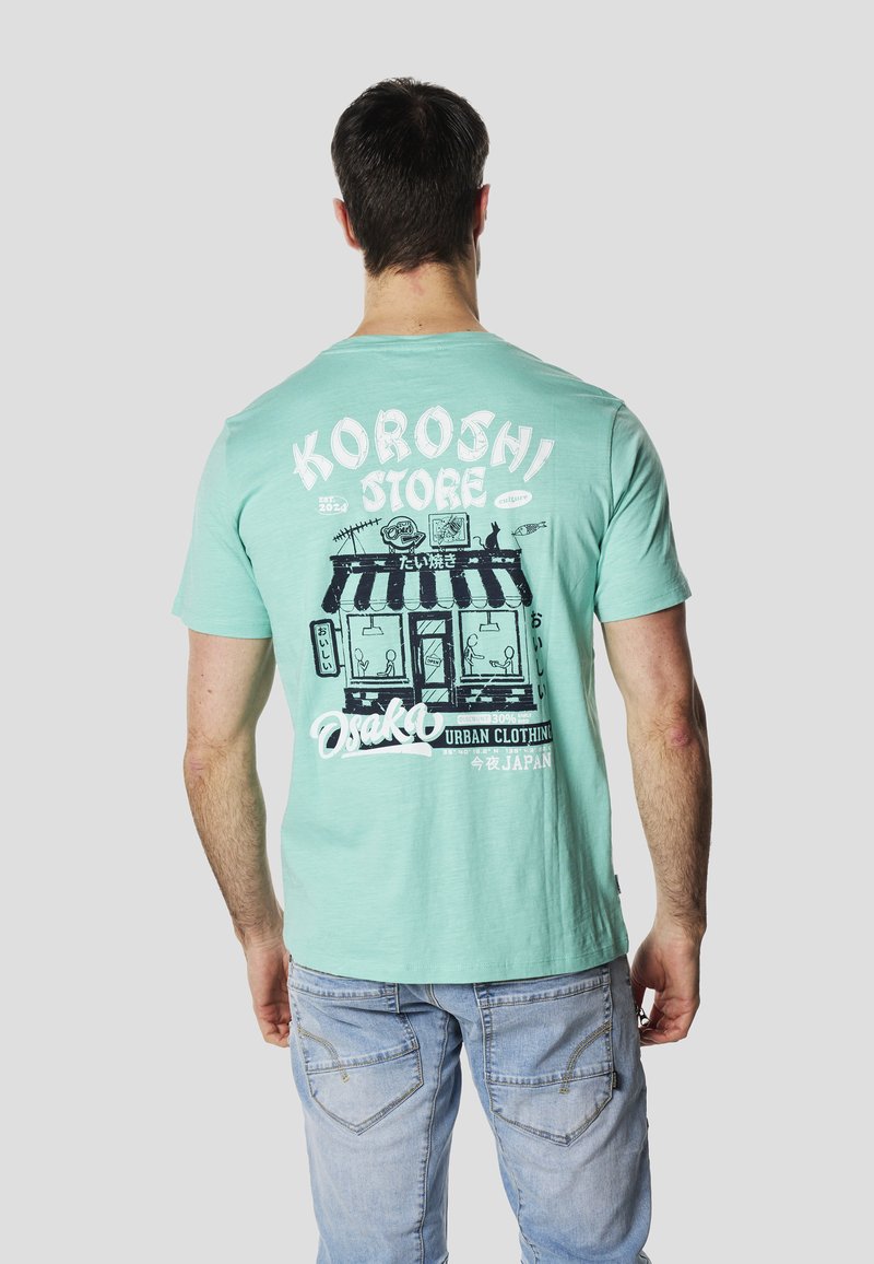 Camiseta verde menta con un estampado gráfico que muestra una fachada de tienda y un texto de logo en blanco y negro, manga corta y un ajuste holgado.