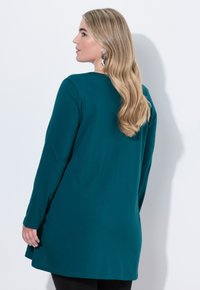 Langärmlige türkisfarbene Tunika aus weichem Stoff mit einem runden Ausschnitt und einem leicht geschwungenen Saum, kombiniert mit schwarzen Hosen.