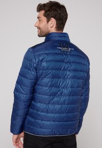 Giacca puffa blu, caratterizzata da motivi a righe orizzontali, collo alto e marchio nero sul retro. Tessuto testurizzato, leggero e resistente all'acqua.