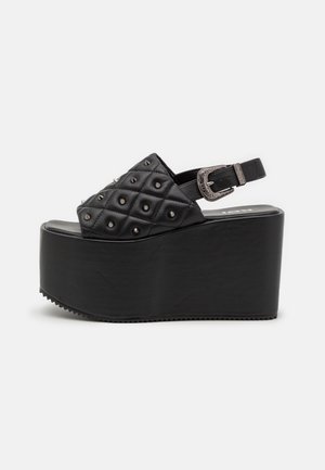 Czarne sandały na platformie mają pikowaną górę z syntetycznego materiału z ćwiekami, regulowany pasek na kostkę oraz grubą podeszwę na koturnie. Teksturowana gumowa podeszwa.