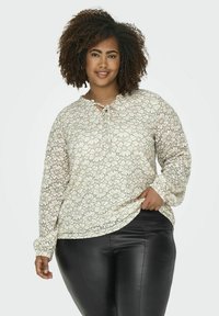Blusa floreale di crema chiara con maniche lunghe e scollatura con nodo, caratterizzata da un'overlay in pizzo. Indossata con pantaloni neri attillati.