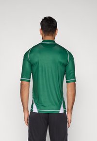 Zielona sportowa koszulka z krótkim rękawem z białymi akcentami po bokach i na ramionach, wykonana z gładkiego, lekkiego materiału; posiada klasyczny kołnierzyk i dopasowany krój.