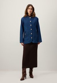 Tmavomodrá rifľová bunda s okrúhlym golierom, zlatými gombíkmi, dvoma bočnými vreckami a voľným strihom, kombinovaná s hnedou sukňou dĺžky po členky.