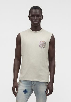Jeune homme à la peau foncée portant un débardeur beige avec un petit motif sur la poitrine et un jean bleu clair avec des patches.