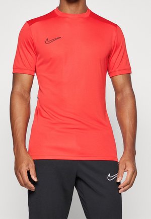 Mann trägt ein rotes kurzärmliges Sportshirt mit schwarzem Nike-Swoosh und schwarze Shorts mit weißem Nike-Logo.