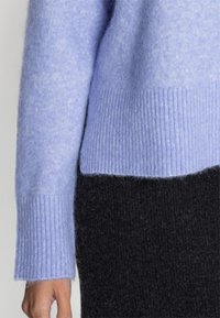 Jasno fioletowy sweter z dzianiny z ribbowymi mankietami, z miękkim, teksturowanym wykończeniem. Połączony z ciemnym czarnym materiałem poniżej.