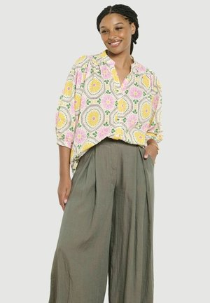 Kobieta z warkoczami, ubrana w luźną kwiecistą bluzkę z żółtymi i różowymi kwiatami oraz szerokie oliwkowozielone spodnie, stojąca z jedną ręką w kieszeni.