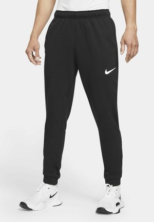 Mężczyzna w czarnych spodniach dresowych Nike z elastycznym pasem i białych butach do biegania Nike, stojący na jednolitym tle.