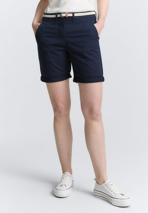 Pantalones cortos azul marino con dobladillos enrollados, que cuentan con bolsillos delanteros y un cinturón tejido de color claro. Combinados con zapatillas blancas.