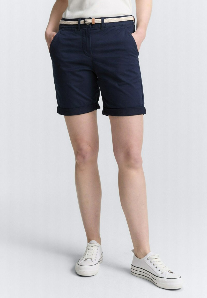 Marineblaue Shorts mit umgeschlagenen Säumen, ausgestattet mit Vordertaschen und einem hellen gewebten Gürtel. Kombiniert mit weißen Sneakers.