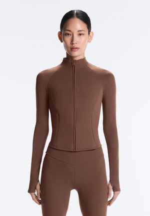 Vrouw draagt een nauwsluitende bruine lange mouwen top met ritssluiting en bijpassende hoog getailleerde leggings, staand tegen een effen witte achtergrond.