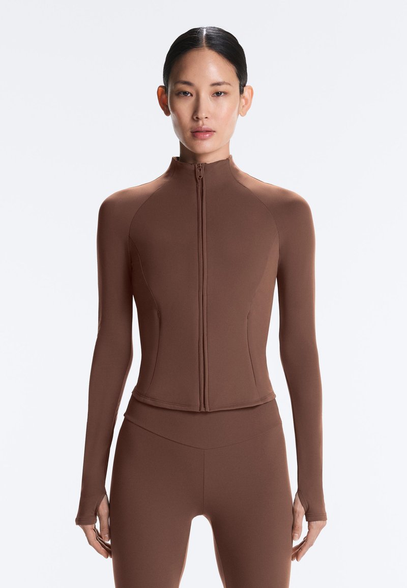 Frau trägt ein eng anliegendes braunes Langarm-Zip-up-Top und passende hoch taillierte Leggings, steht vor einem schlichten weißen Hintergrund.