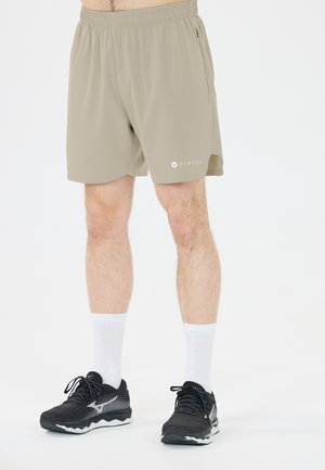 Mann trägt beige Sportshorts mit elastischem Bund, weiße Sneakersocken und schwarze Laufschuhe auf weißem Hintergrund.