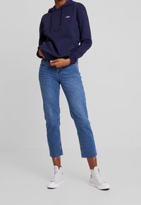 Navy blue hoodie with a front pocket and logo, paired with light blue cropped jeans and white sneakers. Smooth fabric and relaxed fit.