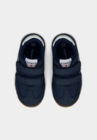 Παιδικά παπούτσια σε ναυτικό μπλε με λουράκια Velcro, συνθετικό επάνω μέρος, λαστιχένια σόλα και λευκές λεπτομέρειες με το όνομα της μάρκας "TOMMY" στη γλώσσα.