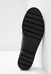 Semelle de botte noire avec des rainures horizontales profondes et « Tamaris 37 » embossé au centre, inclinée contre un fond d'angle blanc.