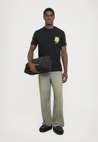 Young man wearing black SpongeBob t-shirt, faded green jeans, holding a black folded bag against a plain white background.