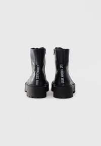 Par de botas de tobillo de cuero negro texturizado con suelas gruesas, vistas desde atrás, con la marca vertical blanca de Steve Madden.