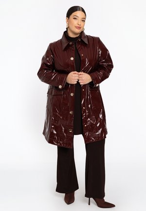 Femme portant un manteau boutonné en bordeaux sombre et brillant sur un pantalon et un haut noirs, se tenant les mains jointes devant un fond blanc.