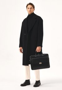 Abrigo de lana negro, cuello de tortuga negro, pantalones blancos y botas de cuero negras. Lleva un elegante maletín de cuero negro con detalles plateados.