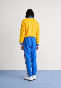 Chándal con una parte superior amarilla con rayas verticales y un cuello azul, combinado con pantalones azules con rayas laterales amarillas y puños elásticos.