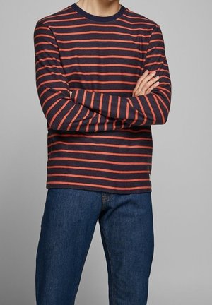 Persona con camiseta de manga larga a rayas azul marino y naranja con los brazos cruzados y jeans azules frente a un fondo neutro.