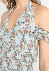 Ljusblå blommig klänning med halterneck och design med kalla axlar. Fina volanger och intrikata blommönster med smockad byst.