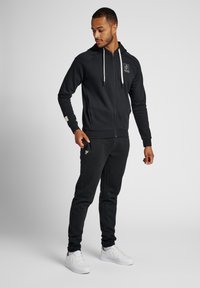 Schwarze Zip-Hoodie mit einem weißen Logo, ausgestattet mit einer Kapuze mit Kordelzug und gerippten Bündchen, kombiniert mit schmal geschnittenen schwarzen Jogginghosen und weißen Sneakers.