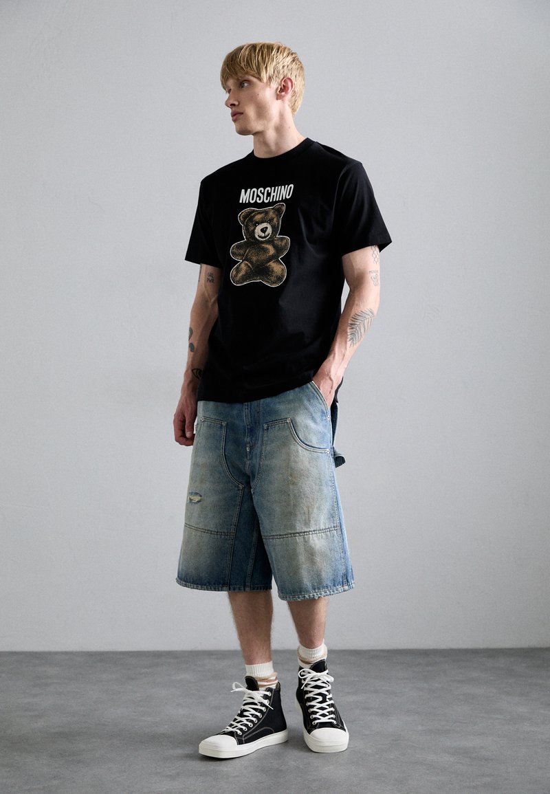 Sort t-shirt med grafik af en bjørn og "MOSCHINO" tekst, parret med lyse denimshorts. Low-top sorte sneakers fuldender looket.