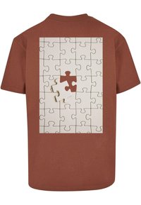 Brun T-shirt med et trykt puslespilsdesign på ryggen, med hvide og tan puslespilsstykker mod en lys baggrund.
