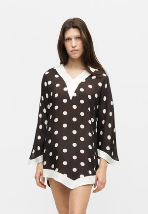 Noor naine pikkade pruuni juustega kannab musta valgete täppidega tuunikat valgete ääristega, seisab lihtsa heleda taustaga.