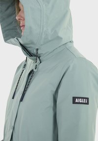 Veste imperméable verte claire avec une grande capuche, des cordons de serrage ajustables et un logo cousu sur la manche gauche. Texture du tissu lisse.