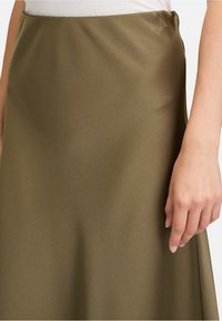 Olivgrüner Satinhrock mit glatter Textur, fließendem Design und dezentem Schimmer. Eine Hand ruht sanft auf dem Stoff und betont dessen Weichheit.