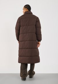 Hnedý puff coat s vysokým golierom, s horizontálnymi prešívanými sekciami, dlhými rukávmi a s dĺžkou do polovice lýtok. Nosené s tmavými nohavicami.