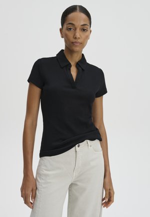 Vrouw met donker haar in een zwart polohemd met korte mouwen en lichte broek, staand tegen een effen lichte achtergrond.