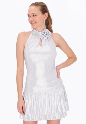 Vrouw draagt een glanzende zilveren halterjurk met een keyhole-halslijn en een stoffen bloem, glimlachend, met lange oorbellen en hand op heup.