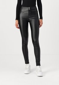 Zwarte, high-waisted nepleren broeken met twee voorzakken en zichtbare stikdetails. Strak ontwerp, gecombineerd met witte sneakers.