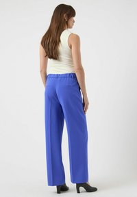 Blå vidbukser med elastisk midje, sidelommer og jevn tekstur, kombinert med en kremfarget ermeløs topp og svarte hæler.