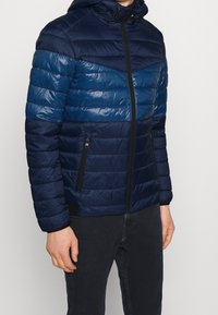 Marinblå pufferjacka med horisontellt quiltade sektioner, huva och svarta dragkedjefickor. Tillverkad av lättviktig, glänsande material.