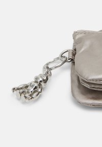 Silberner, metallischer Schlüsselanhänger mit einer Bärenfigur, die an einem hellgrauen, strukturierten Beutel befestigt ist. Der Beutel hat einen Druckverschluss und einen dezenten Glanz.