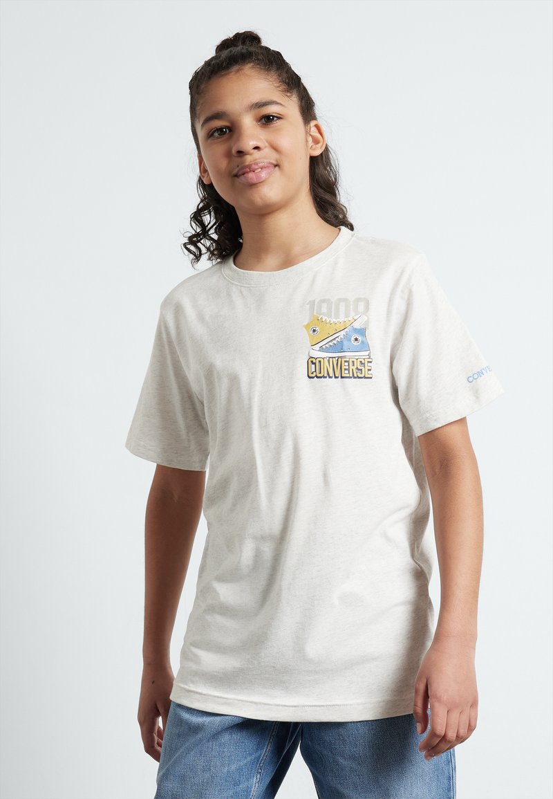 Ljusbeige bomullt-shirt med en grafik av Converse-skor i blått och gult med varumärkesnamnet. Korta ärmar och rund halsringning.