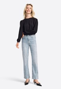 Top negro de manga larga con escote fruncido, jeans de tiro alto en azul claro y tacones negros de punta, de pie contra un fondo blanco.