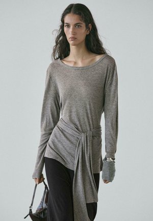 Maglia grigia a maniche lunghe in tessuto morbido con scollo ampio, dettaglio con nodo in vita e accenti testurizzati sulle maniche. Indossata con pantaloni scuri e una borsa.