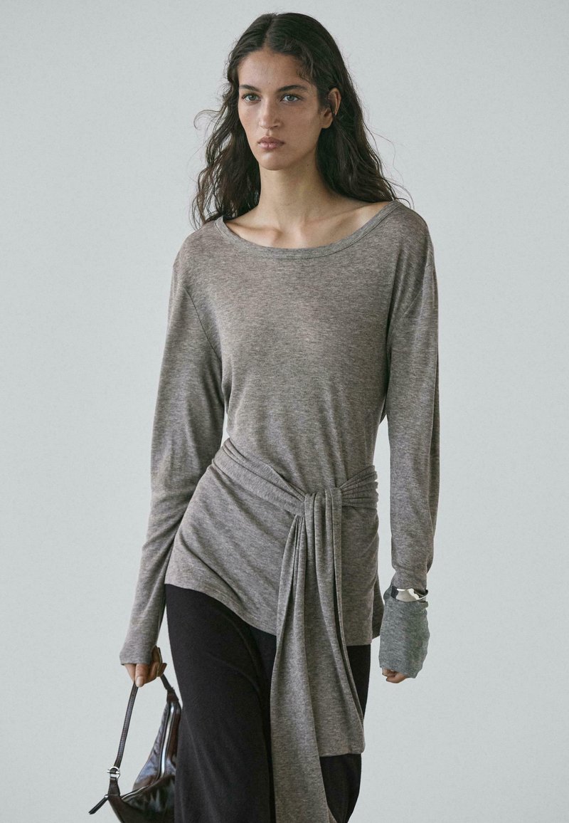 Szary top z długim rękawem wykonany z miękkiego materiału, z szerokim dekoltem, detalem z supłem w talii oraz teksturowanymi akcentami na rękawach. Noszony z ciemnymi spodniami i torbą.