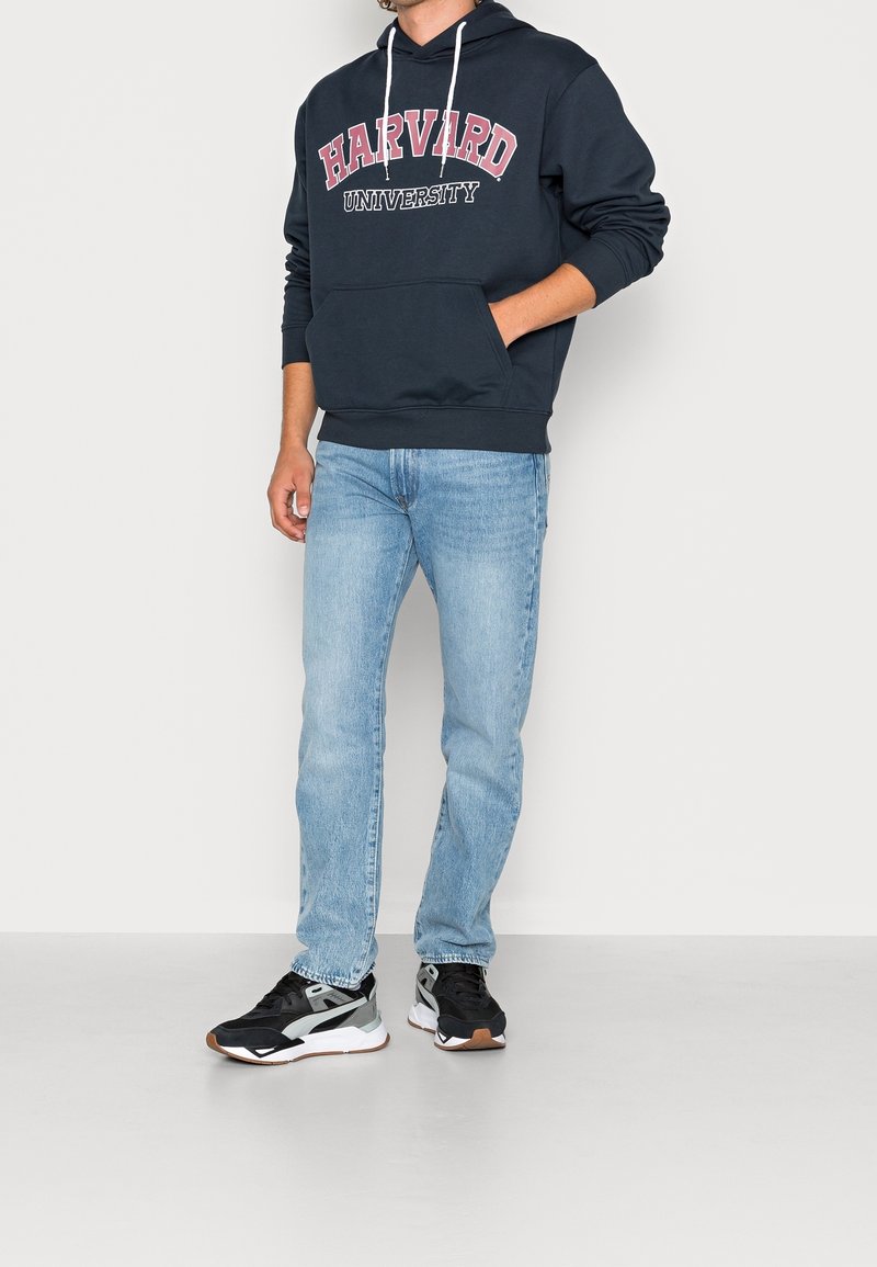 Personne portant un sweat à capuche bleu marine de l'Université de Harvard, un jean bleu clair et des baskets noires et blanches, debout devant un fond blanc uni.