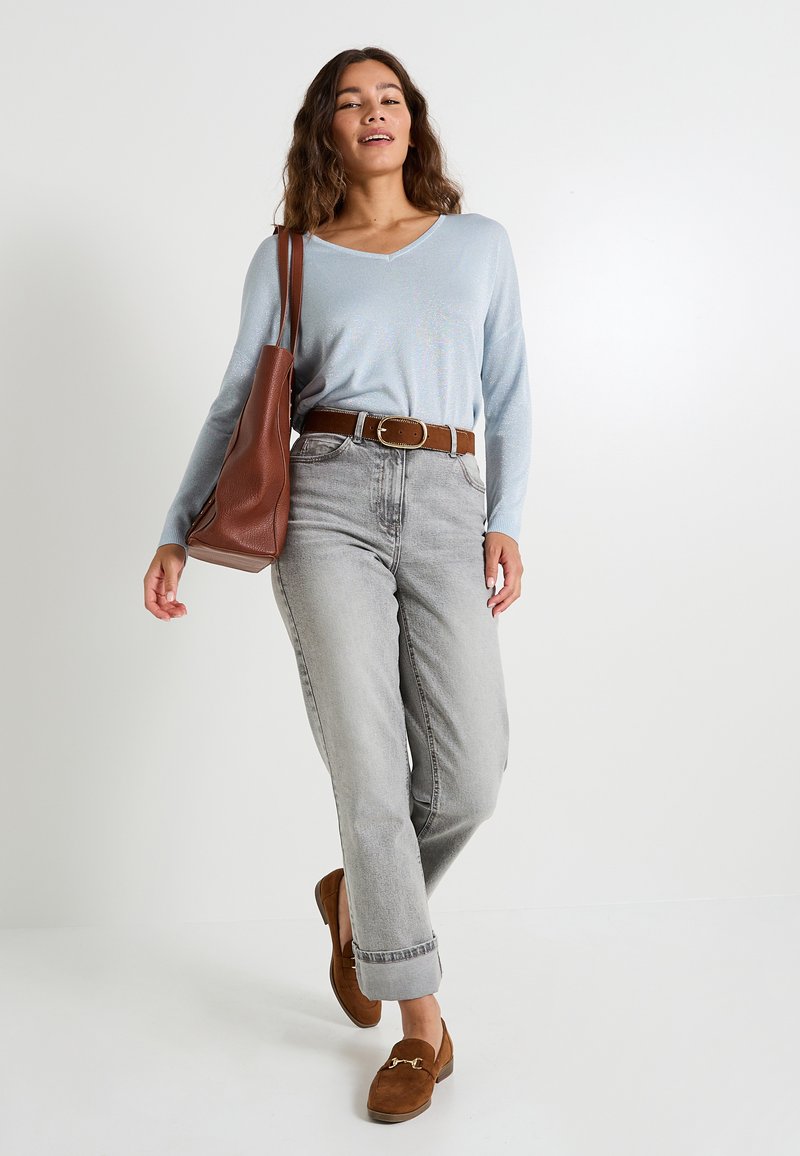 Haut à manches longues bleu clair, pantalon taille haute gris, ceinture en cuir marron, mocassins en cuir suédé marron et un sac fourre-tout assorti, debout contre un fond blanc.