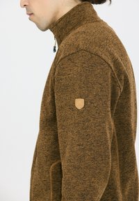 Giacca in pile marrone, textured, con colletto alto, chiusura a zip e un piccolo emblema beige sulla spalla sinistra.