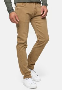 Vaqueros slim-fit de color marrón claro, fabricados en denim suave, con bolsillos delanteros, pasadores para cinturón y sutiles detalles de costura. Combinados con zapatillas blancas.