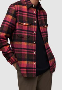 Koszula flanelowa w kratę w kolorze różowym, pomarańczowym, czarnym i brązowym. Posiada dwie kieszenie na piersi i białe guziki. Miękka tekstura, luźny krój.